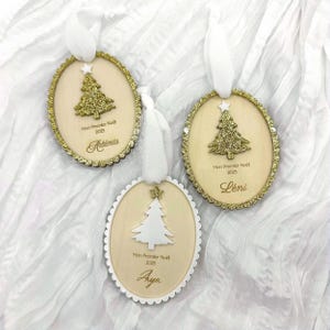 K&ouml;nnte beinhalten: Drei ovale Weihnachtsanh&auml;nger aus Holz mit wei&szlig;en B&auml;ndern. Zwei zeigen goldene Glitzerb&auml;ume mit Sternen, einer einen wei&szlig;en Baum mit Stern. Jeder Anh&auml;nger ist mit Mon Premier No&euml;l 2025 und den Namen Art&eacute;mis, L&eacute;ni oder Aya beschriftet.