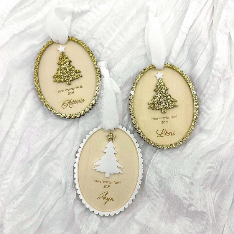 K&ouml;nnte beinhalten: Drei ovale Weihnachtsanh&auml;nger aus Holz mit wei&szlig;en B&auml;ndern. Zwei zeigen goldene Glitzerb&auml;ume mit Sternen, einer einen wei&szlig;en Baum mit Stern. Jeder Anh&auml;nger ist mit Mon Premier No&euml;l 2025 und den Namen Art&eacute;mis, L&eacute;ni oder Aya beschriftet.