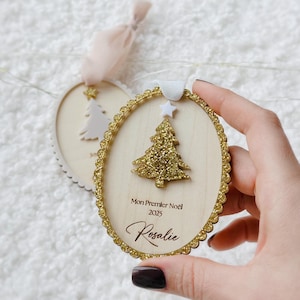 K&ouml;nnte beinhalten: Ein ovaler Weihnachtsbaumschmuck aus Holz mit einem goldglitzernden Weihnachtsbaum, einem wei&szlig;en Stern als Spitze und einem gewellten Goldglitterrand. Der Text lautet Mon Premier No&euml;l 2025 und der Name Rosalie mit wei&szlig;er Schleife.