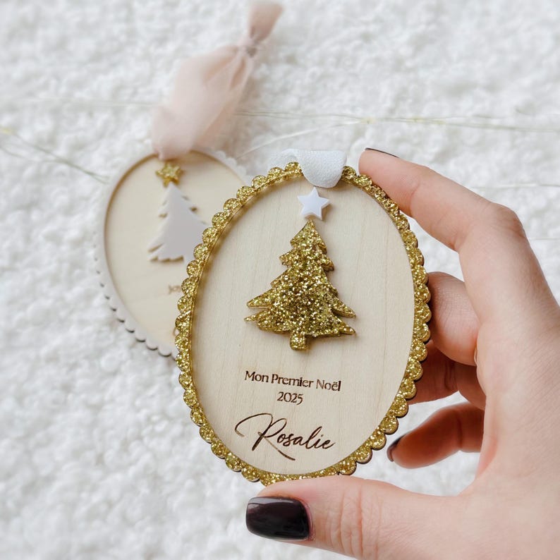 K&ouml;nnte beinhalten: Ein ovaler Weihnachtsbaumschmuck aus Holz mit einem goldglitzernden Weihnachtsbaum, einem wei&szlig;en Stern als Spitze und einem gewellten Goldglitterrand. Der Text lautet Mon Premier No&euml;l 2025 und der Name Rosalie mit wei&szlig;er Schleife.