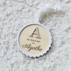 Könnte beinhalten: Ein runder Weihnachtsbaumschmuck aus Holz mit einem weißen, gewellten Rand. Der Schmuck zeigt einen großen Buchstaben "A", den Text "MON PREMIER NOËL 2025" und den Namen "Agathe". Eine weiße Stoffschlaufe ist zum Aufhängen befestigt.