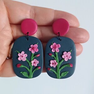 May include: A pair of dark blue statement earrings with pink and burgundy floral accents. The earrings are made of polymer clay and have a unique, handmade design.