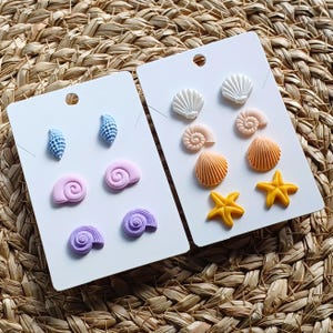 Könnte beinhalten: Zwei Sets von Ohrringen mit Muschel-Motiv auf weißem Karton. Das erste Set zeigt lila, rosa und blaue Muscheln. Das zweite Set zeigt weiße, orange und gelbe Muscheln mit Seesternen.