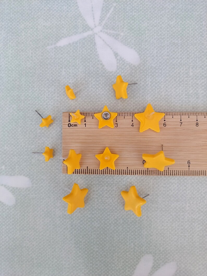 Three pairs of yellow star shaped stud earrings in small, medium and large with a wooden ruler and green background with white dragonflies