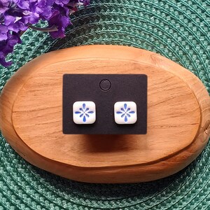 May include: A pair of square stud earrings with a white base and blue floral design. The earrings are displayed on a black card, set on a wooden oval-shaped tray. The background is a green textured surface.