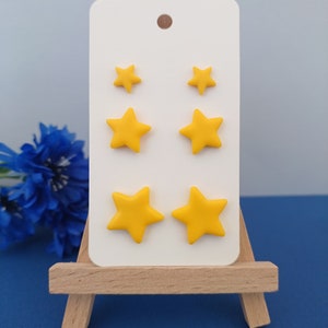 Three pairs of yellow star shaped stud earrings in small, medium and large on a white display card with a grey and blue background and blue flowers