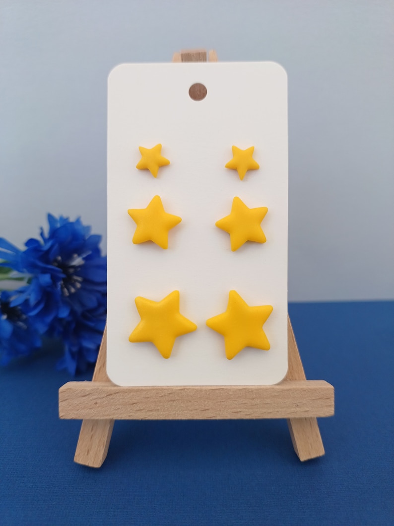 Three pairs of yellow star shaped stud earrings in small, medium and large on a white display card with a grey and blue background and blue flowers