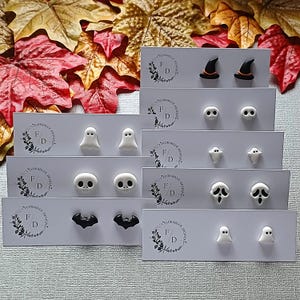 Puede incluir: Una colección de pendientes de botón con temática de Halloween. Los diseños incluyen sombreros de bruja, fantasmas, calaveras y murciélagos. Las tarjetas se presentan sobre un fondo de hojas de otoño. La marca "FLORABLUE DESIGNS" es visible.