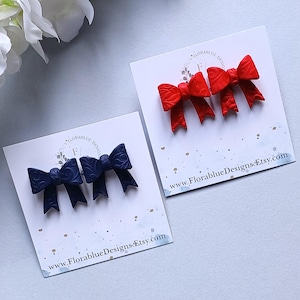 May include: Two sets of bow-shaped hair clips, one in red and the other in navy blue, displayed on white cards. The red bows have a textured surface, while the navy bows have a patterned design. Each card has the website address www.florabluedesigns.etsy.com.