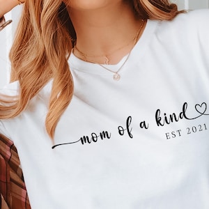 以下が含まれることがあります： 「mom of a kind」という黒い文字が書かれた白いTシャツ。小さなハートマークと「EST 2021」が下に書かれている。