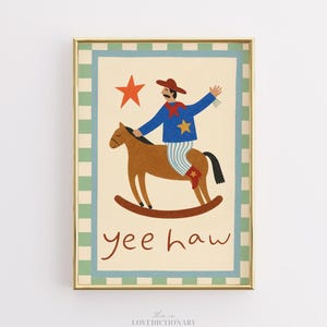 May include: A framed print featuring a cowboy on a rocking horse. The cowboy wears a blue jacket, red hat, and striped pants. A red star is above the cowboy, and the words "yee haw" are below. The frame has a gold finish.