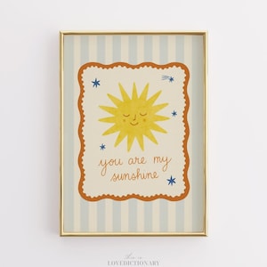 Puede incluir: Un estampado enmarcado con un sol amarillo y el texto "you are my sunshine". El estampado tiene un borde crema con un borde festoneado y está sobre un fondo a rayas azules y blancas. El marco es dorado.