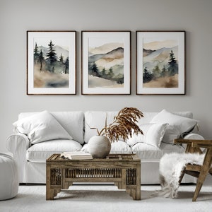 May include: Three framed watercolour landscape prints. Each print depicts a mountain scene with trees in shades of green, brown, and grey. The frames are dark brown. The prints are displayed above a white sofa and wooden coffee table.