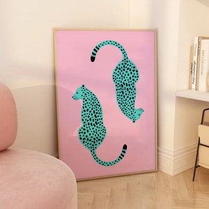 May include: A framed print with a pink background featuring two turquoise cheetahs with black spots. The cheetahs are facing opposite directions and are in a curved shape.