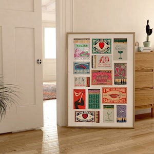 May include: A framed print of vintage matchbox labels with colorful designs and text. The labels feature images of hearts, lips, cocktails, and bicycles. The print is displayed on a wall in a room with a wooden dresser and a potted plant.