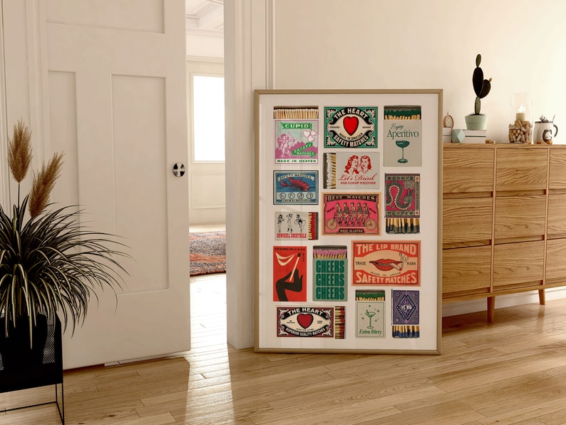 May include: A framed print of vintage matchbox labels with colorful designs and text. The labels feature images of hearts, lips, cocktails, and bicycles. The print is displayed on a wall in a room with a wooden dresser and a potted plant.