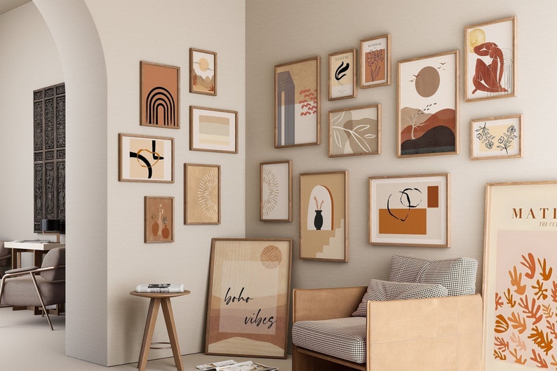 May include: A gallery wall with a variety of framed abstract art prints in shades of brown, beige, and black. The prints feature geometric shapes, lines, and organic forms. The wall is painted a light beige color and the frames are all made of wood.