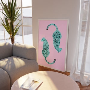 May include: A framed print with a pink background featuring two teal blue cheetahs with black spots. The cheetahs are facing opposite directions and are in a sitting position.