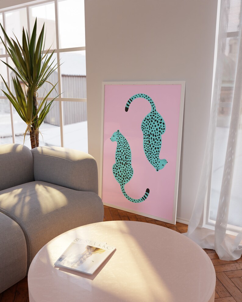 May include: A framed print with a pink background featuring two teal blue cheetahs with black spots. The cheetahs are facing opposite directions and are in a sitting position.