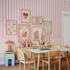May include: A dining room with a pink and white striped wall, adorned with framed art prints. A wooden cabinet holds a pink lamp, books, and a pink magazine rack. A light blue stool and a white table with wooden chairs complete the scene.