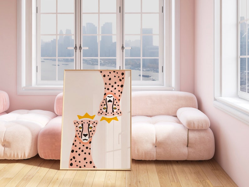 May include: A framed print of two pink cheetahs with black spots. The cheetahs are wearing gold crowns and are looking at the viewer. The print is hanging on a pink wall in a room with a pink sofa and windows.