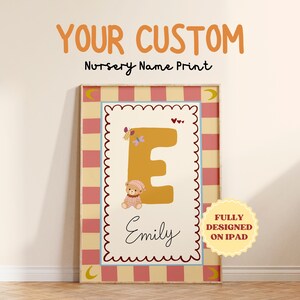 May include: Custom nursery name print featuring a large gold letter "E" with a teddy bear illustration. The print includes the name "Emily" in cursive, butterflies, and hearts. The background is a pink and cream plaid pattern.
