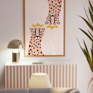 May include: A framed print of two pink cheetahs with black spots, one upside down, both wearing gold crowns. The background is a light pink with a white marble pattern.
