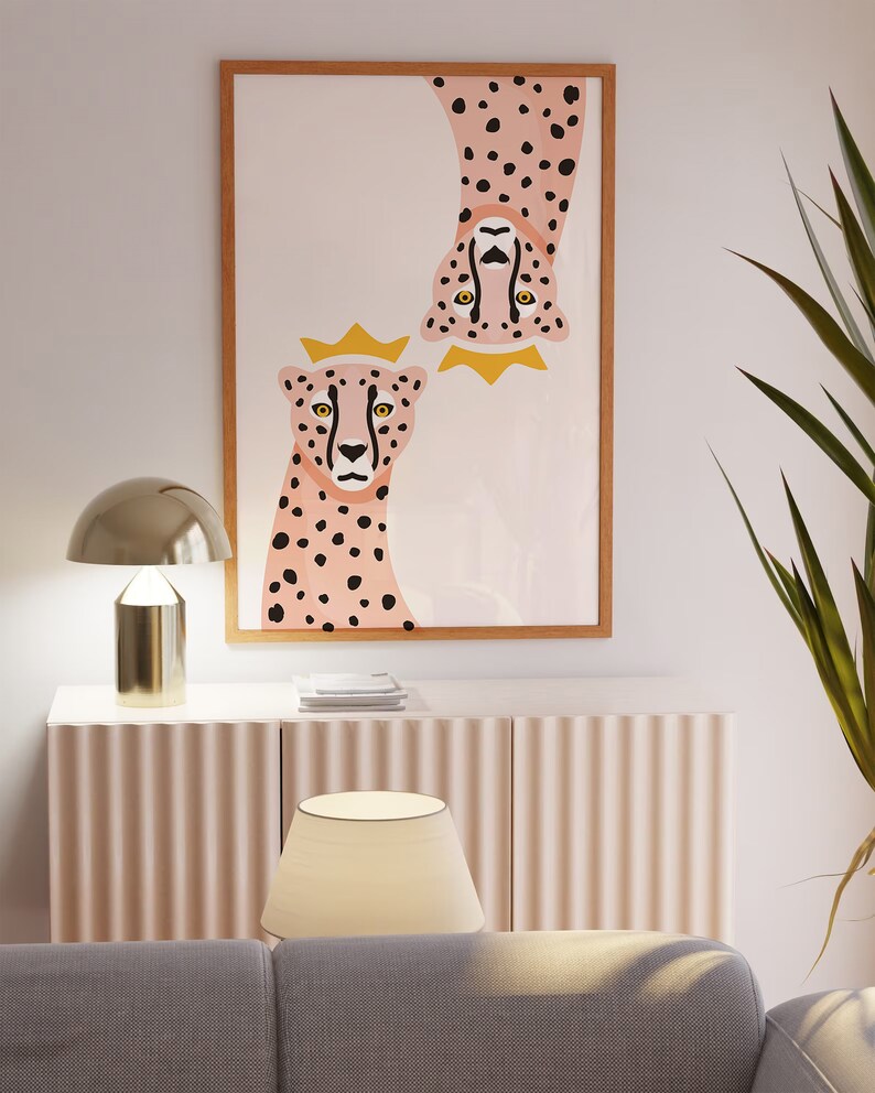 May include: A framed print of two pink cheetahs with black spots, one upside down, both wearing gold crowns. The background is a light pink with a white marble pattern.