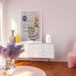 May include: A white framed print with a vintage illustration of a woman in a green swimsuit sitting in a glass of gin and tonic. The print is hanging on a white wall above a white dresser with three drawers. The text on the print reads "Guest Check" and "669906".