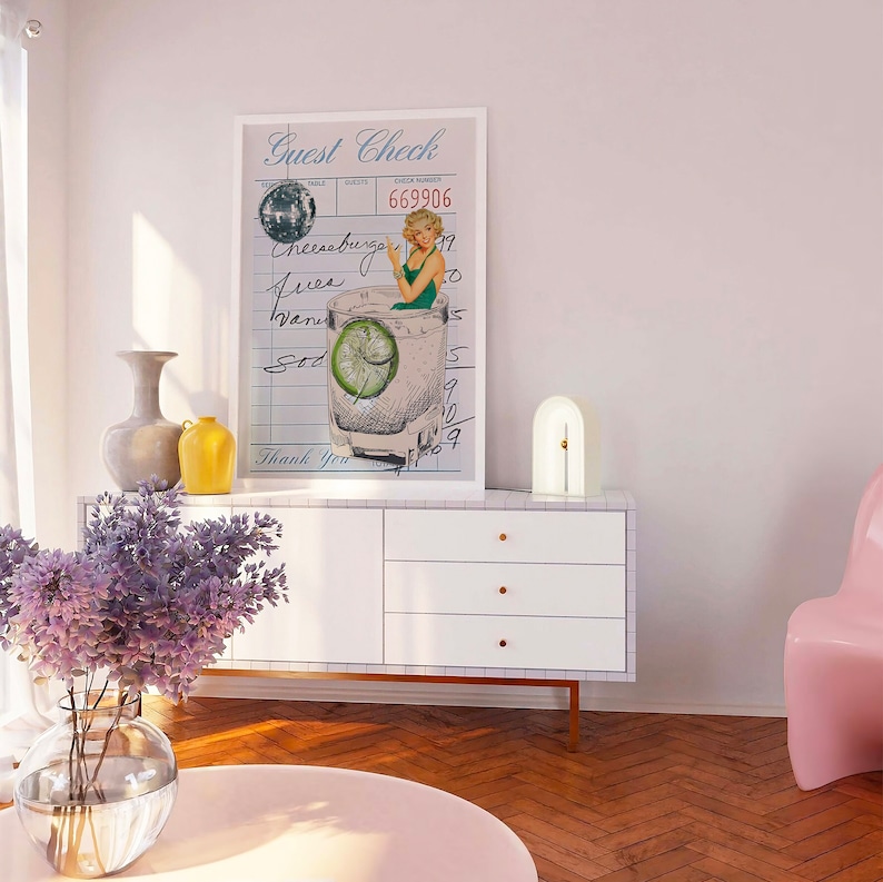 May include: A white framed print with a vintage illustration of a woman in a green swimsuit sitting in a glass of gin and tonic. The print is hanging on a white wall above a white dresser with three drawers. The text on the print reads "Guest Check" and "669906".