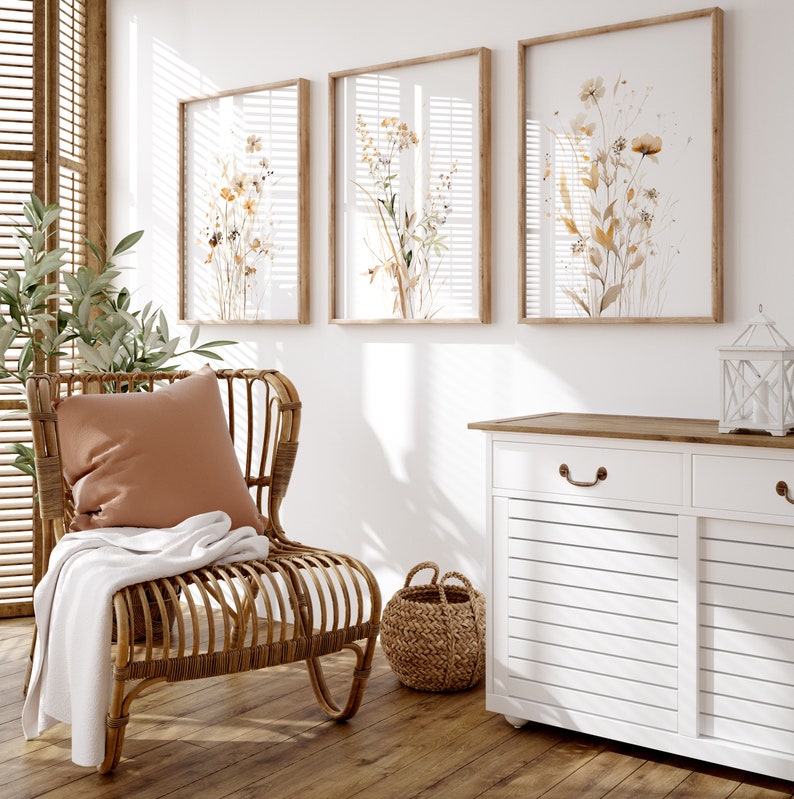 May include: A white cabinet with two drawers and two doors, each with a horizontal slat design. The cabinet has a wooden top and brass handles. A wicker basket sits on the floor in front of the cabinet. A rattan chair with a brown cushion and a white throw blanket sits in front of the cabinet. Three framed botanical prints with watercolor illustrations of flowers and leaves hang on the wall behind the cabinet.