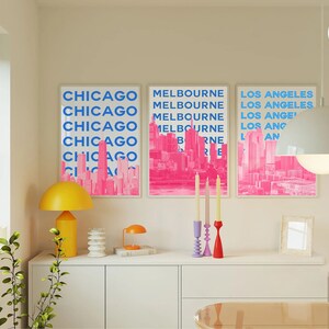 May include: Three pink and blue city skyline prints with the names of the cities in blue text: Chicago, Melbourne, and Los Angeles. The prints are framed and hanging on a white wall above a white dresser.