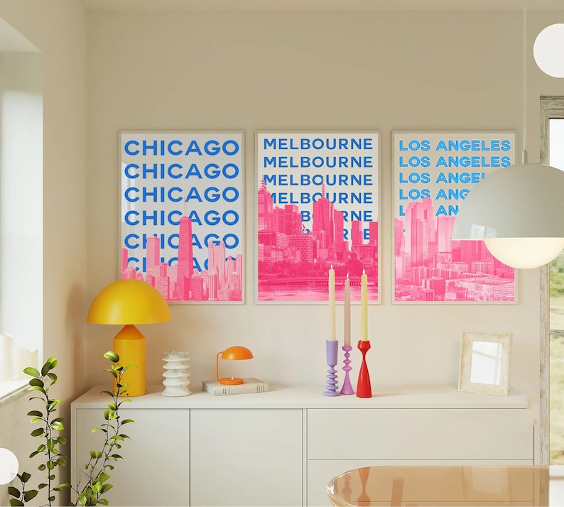 May include: Three pink and blue city skyline prints with the names of the cities in blue text: Chicago, Melbourne, and Los Angeles. The prints are framed and hanging on a white wall above a white dresser.