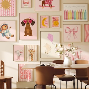 May include: A collection of pink and white framed illustrations with various designs, including a sun, a dog, a giraffe, a bunny, a bow, and a castle. The illustrations are hung on a white wall with a wooden chair and a table in the foreground.