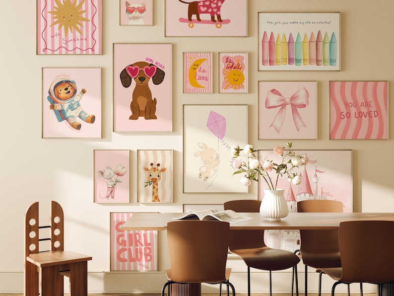 May include: A collection of pink and white framed illustrations with various designs, including a sun, a dog, a giraffe, a bunny, a bow, and a castle. The illustrations are hung on a white wall with a wooden chair and a table in the foreground.