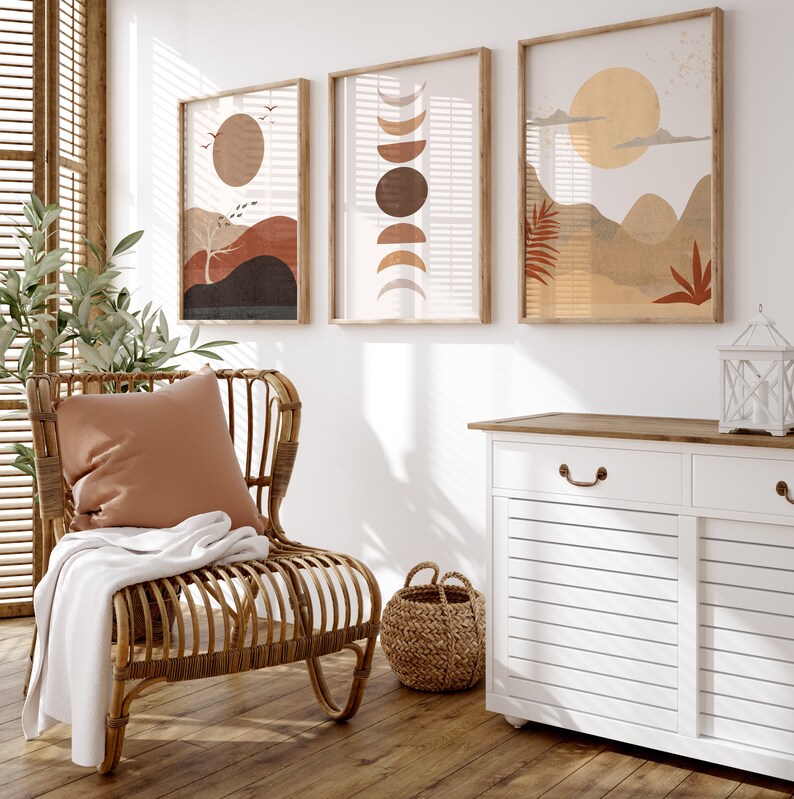 May include: A white cabinet with two drawers and two doors, a wicker chair with a brown throw blanket and a pillow, a woven basket, and three framed abstract art prints with brown, orange, and yellow colors.