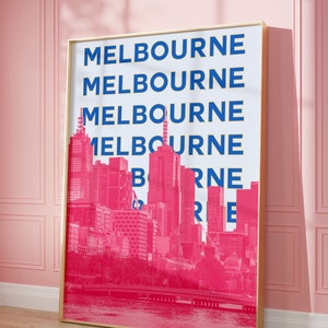 May include: A gold framed print with a white background and the word "MELBOURNE" repeated in blue and yellow lettering. The background image is a pink silhouette of the Melbourne skyline.