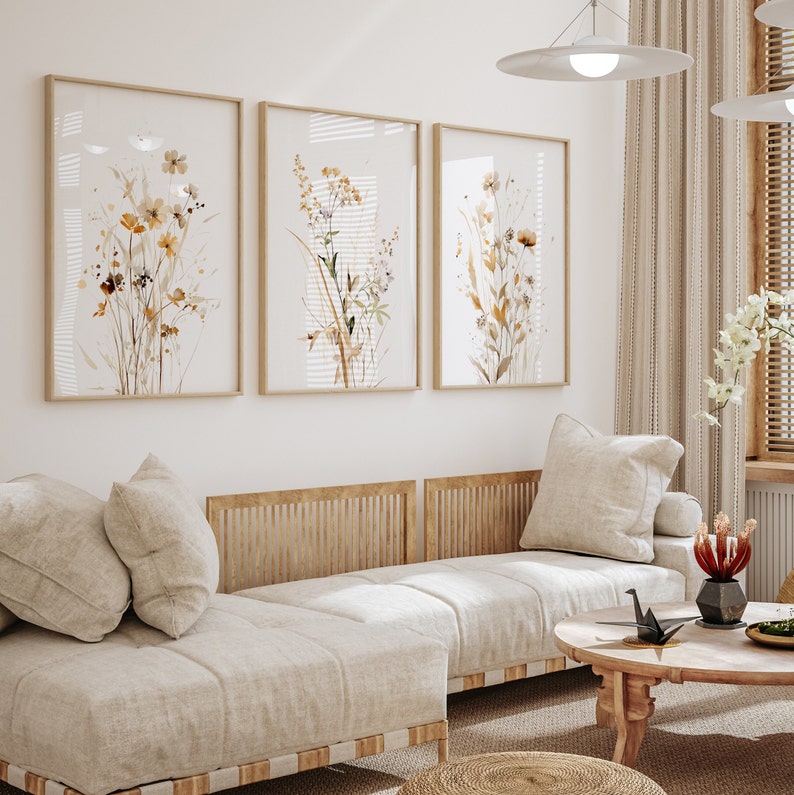 May include: Three framed watercolor prints of dried flowers in shades of brown and beige, hanging on a white wall above a light beige couch with wooden slats.