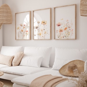 May include: Three framed watercolor prints of delicate peach and yellow flowers with green stems and leaves on a white background. The prints are hung on a white wall above a white couch with pillows and a throw blanket.