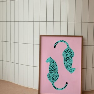 May include: A framed print with a pink background featuring two teal blue leopards with black spots. The leopards are facing opposite directions, one is curled up and the other is sitting upright.