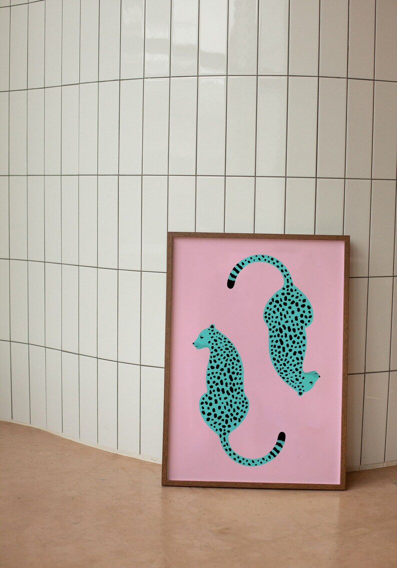 May include: A framed print with a pink background featuring two teal blue leopards with black spots. The leopards are facing opposite directions, one is curled up and the other is sitting upright.