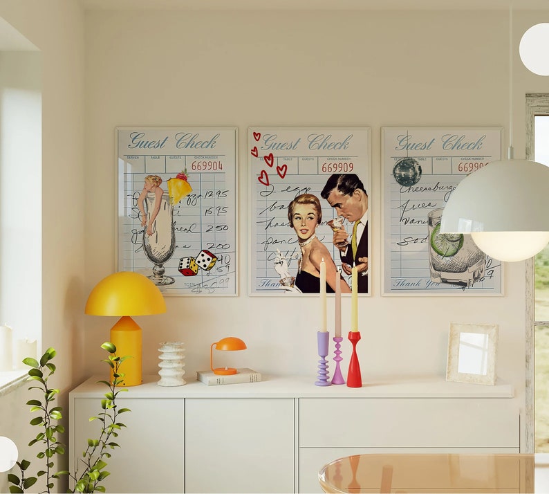 May include: Three vintage guest check style prints with illustrations of cocktails and people. The prints are framed and hung on a white wall above a white shelf with a yellow lamp, a stack of books, and a candle holder with three candles.