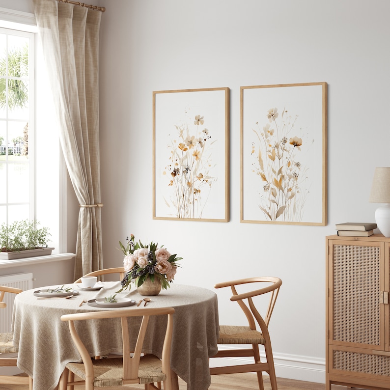 May include: Two framed watercolor prints of brown and beige wildflowers on a white background. The prints are hanging on a white wall above a wooden dining table with a beige tablecloth and two chairs.
