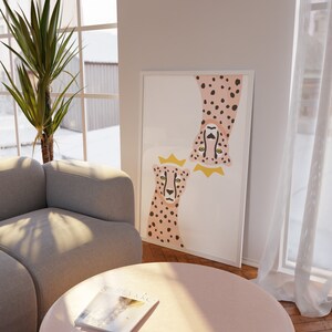 May include: A white framed print with a pink and black cheetah illustration. The cheetah is wearing a gold crown. The print is hanging on a white wall above a light grey sofa and a pink round coffee table.