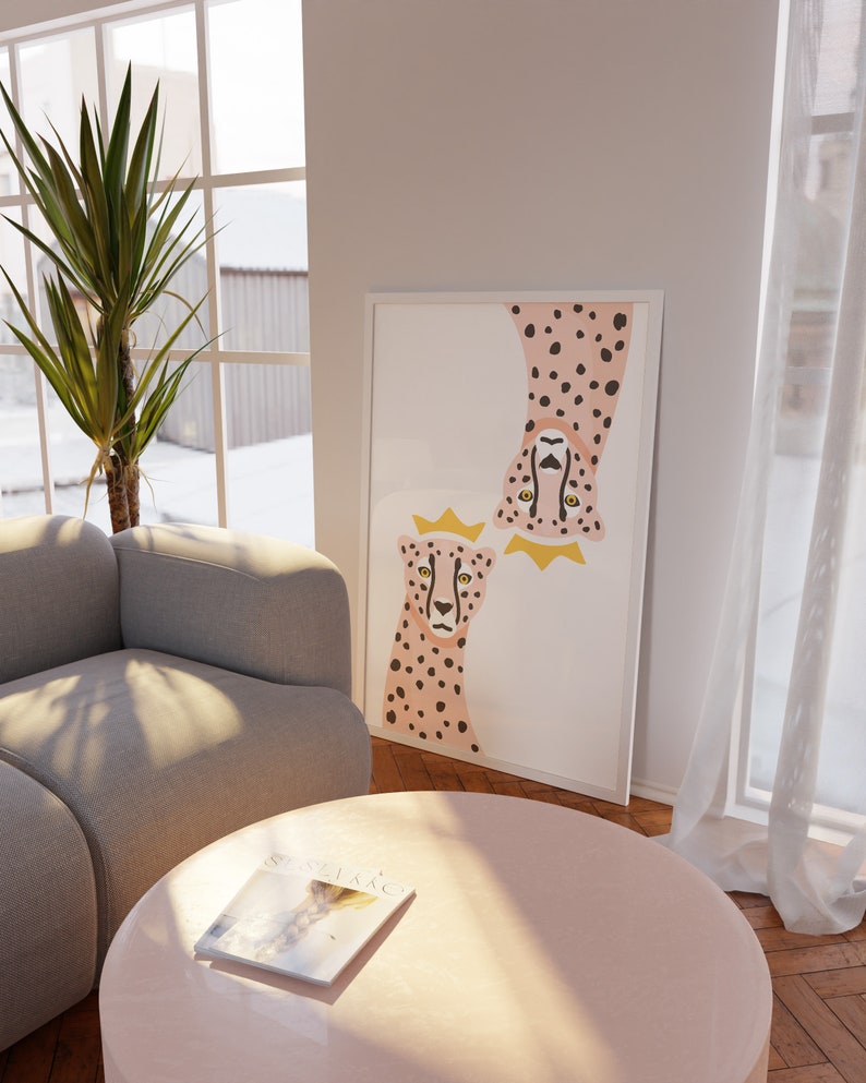 May include: A white framed print with a pink and black cheetah illustration. The cheetah is wearing a gold crown. The print is hanging on a white wall above a light grey sofa and a pink round coffee table.