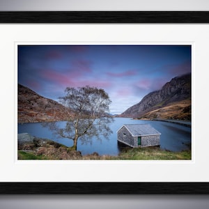 May include: A stone building stands on the edge of a lake, with a tree in the foreground. The sky is a vibrant blue with pink and purple clouds.