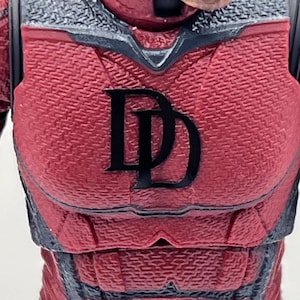 Puede incluir: Una figura de acción roja y gris oscuro de un personaje con un traje texturizado y una máscara con cuernos. El pecho presenta un logotipo negro de doble D. La figura tiene articulaciones visibles en los hombros y los brazos.