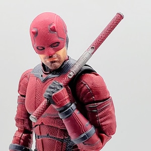 Puede incluir: Figura de acción roja de un superhéroe con un traje texturizado, máscara y guantes, sosteniendo un arma roja y plateada. La figura tiene una expresión decidida y está posada como si estuviera lista para la acción. El disfraz de la figura es detallado.