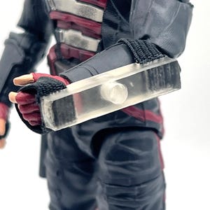 May include: A clear, rectangular accessory with a cylindrical white component is attached to a black sleeve. The accessory is positioned over a red and black glove. The figure is wearing a black and red outfit.