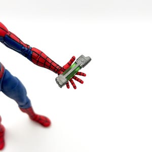 May include: A Spider-Man action figure holding a silver and green device. The figure is in a dynamic pose, with red and blue costume details. The device has a green, glowing interior.